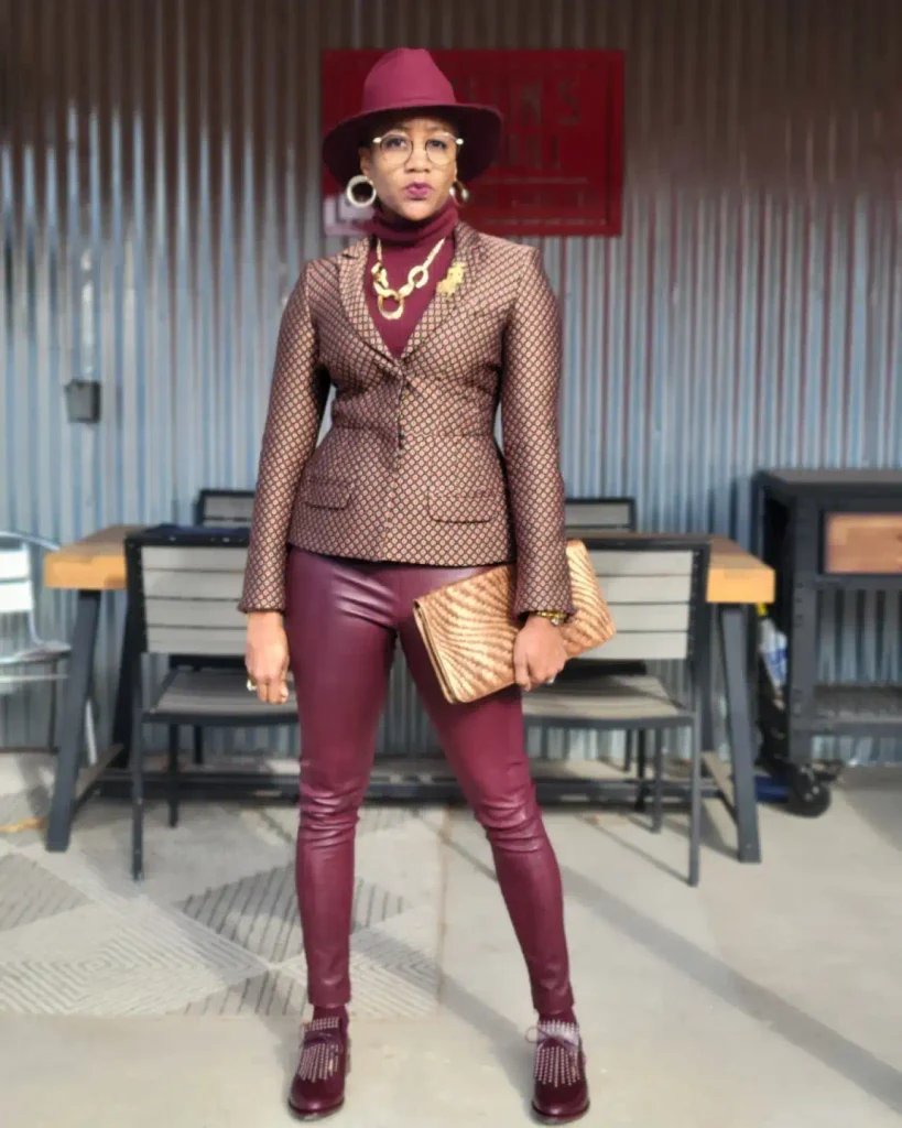 Black woman wearing a patterned tailored blazer with burgundy leather-look pants, matching turtleneck, wide-brim hat, and gold accessories for a sophisticated fall outfit.