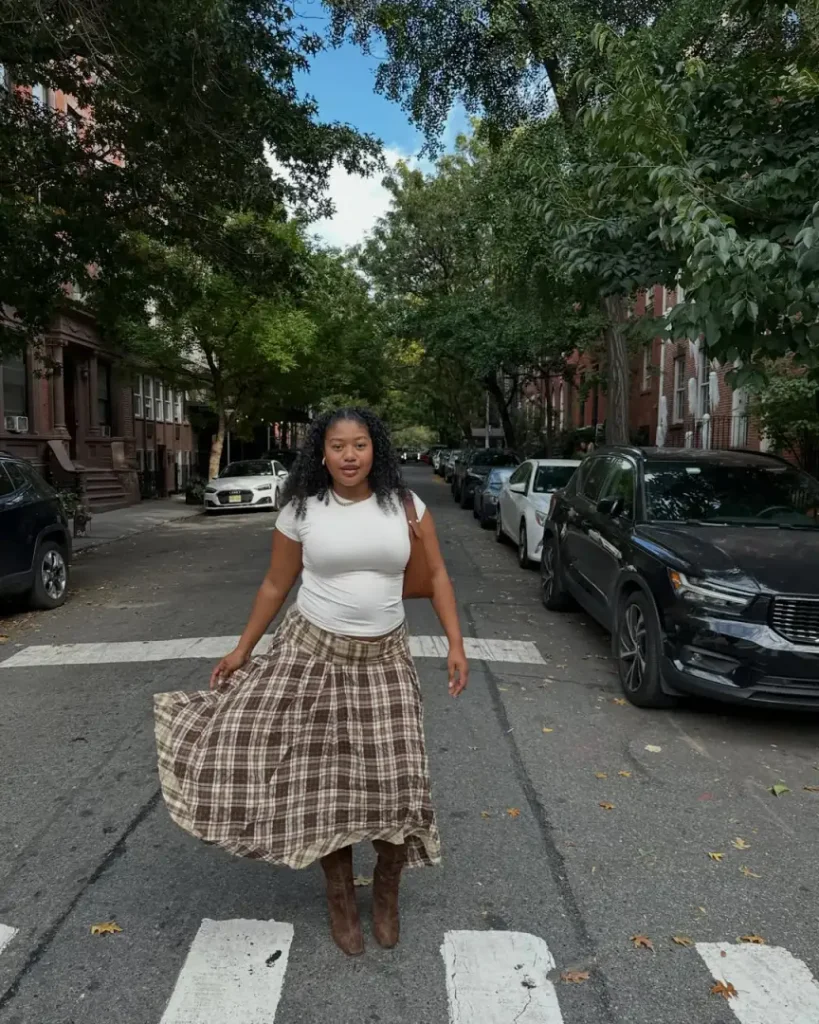Black woman wearing a fitted white T-shirt with a flowing brown plaid maxi skirt, tall suede-look boots, and a brown shoulder bag for fall street style.