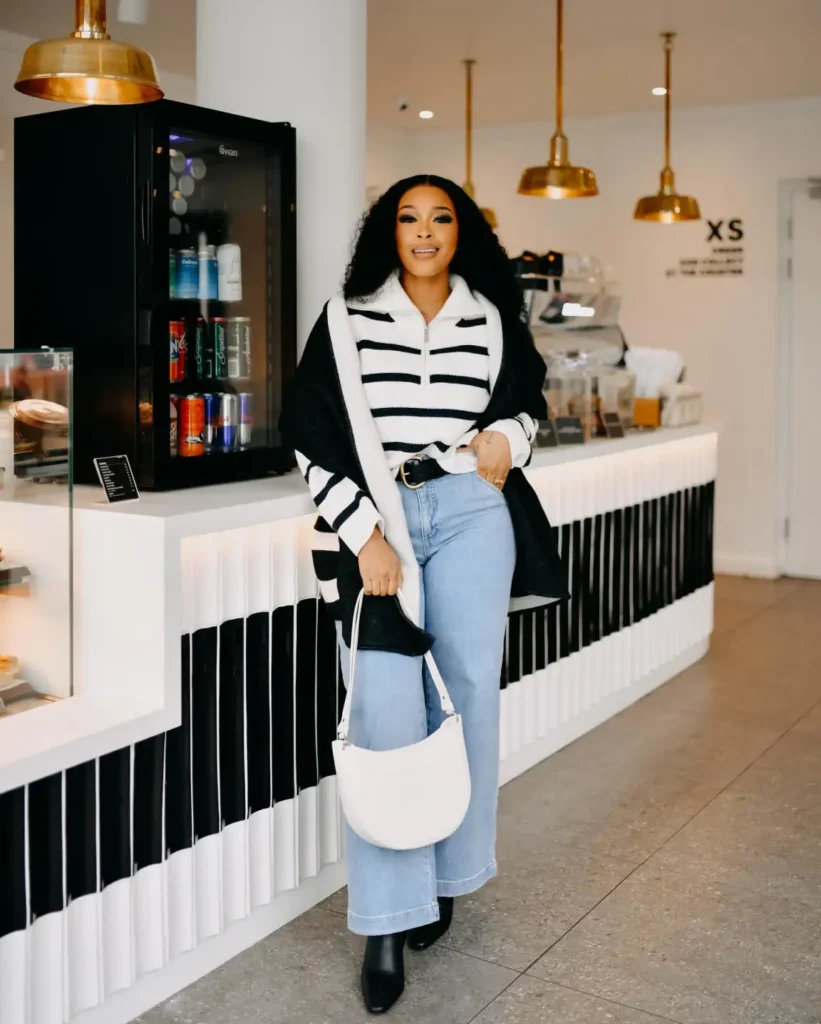Black woman wearing a black-and-white striped quarter-zip sweater with light-wash wide-leg jeans, a black shoulder layer, pointed ankle boots, and a white crescent handbag.