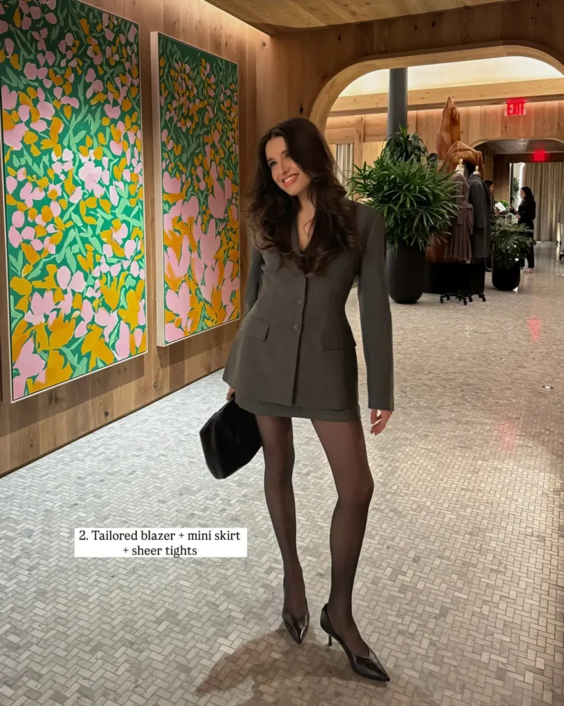 Woman wearing a fitted charcoal blazer with a matching mini skirt, sheer black tights, pointed heels, and a black clutch for a sophisticated fall outfit.