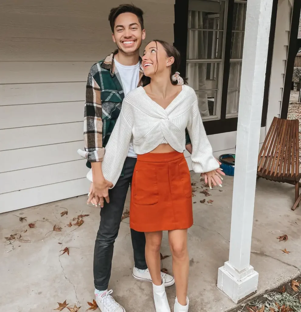 Couple wearing coordinating Thanksgiving outfits with a burnt-orange mini skirt, white twist-front sweater, green plaid overshirt, charcoal jeans, and white footwear.