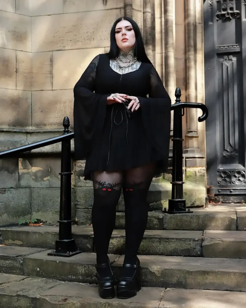 Plus size woman wearing a black Gothic mini dress with dramatic bell sleeves, dark tights, and platform shoes.