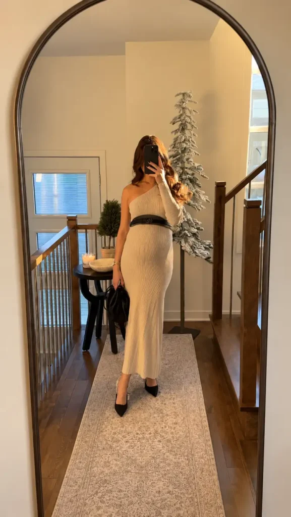 Pregnant woman wearing a cream one-shoulder ribbed knit maxi dress with a black belt, pointed heels, and a black handbag for a stylish Thanksgiving dinner look.