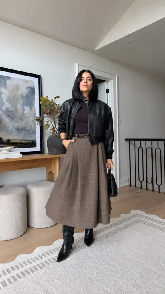 Petite woman wearing a cropped black leather jacket with a brown plaid full midi skirt, black belt, and pointed knee-high boots for fall.