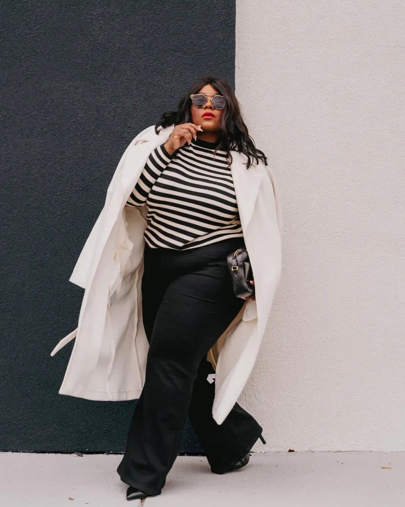 Plus size Black woman wearing a black and cream striped sweater with wide-leg black jeans, a long ivory coat, pointed boots, and sunglasses for a classy fall outfit.