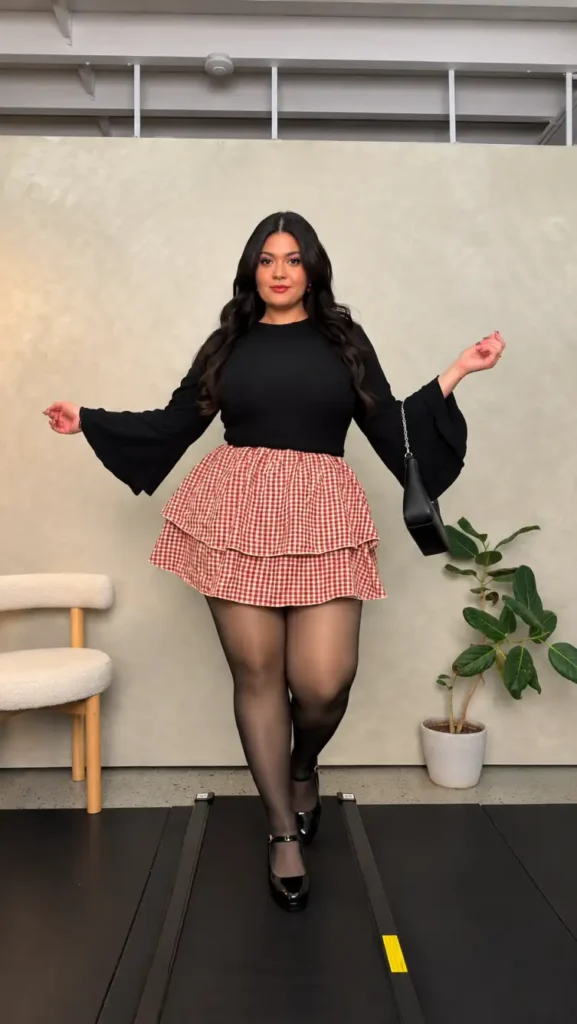 Mid size woman wearing a black bell-sleeve knit top with a red and white gingham tiered mini skirt, sheer black tights, and patent Mary Jane heels.