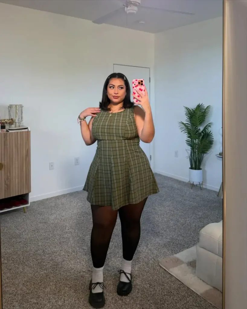 Plus size woman wearing an olive plaid sleeveless mini dress with black tights, white socks, and chunky Mary Jane shoes for fall.