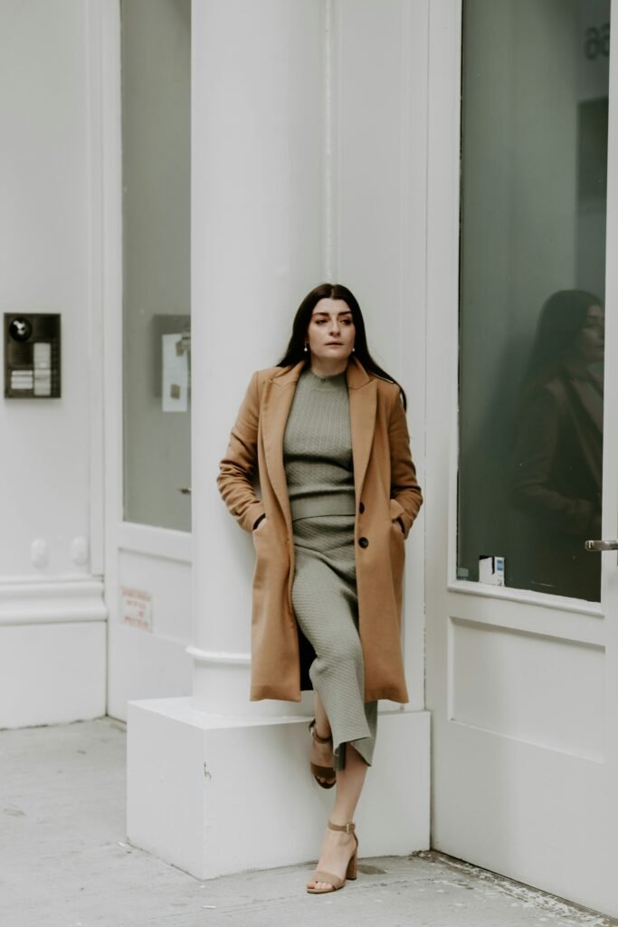 Woman wearing a sage-green ribbed knit midi dress under a long camel coat, paired with nude ankle-strap heels for a classy cold-weather outfit.