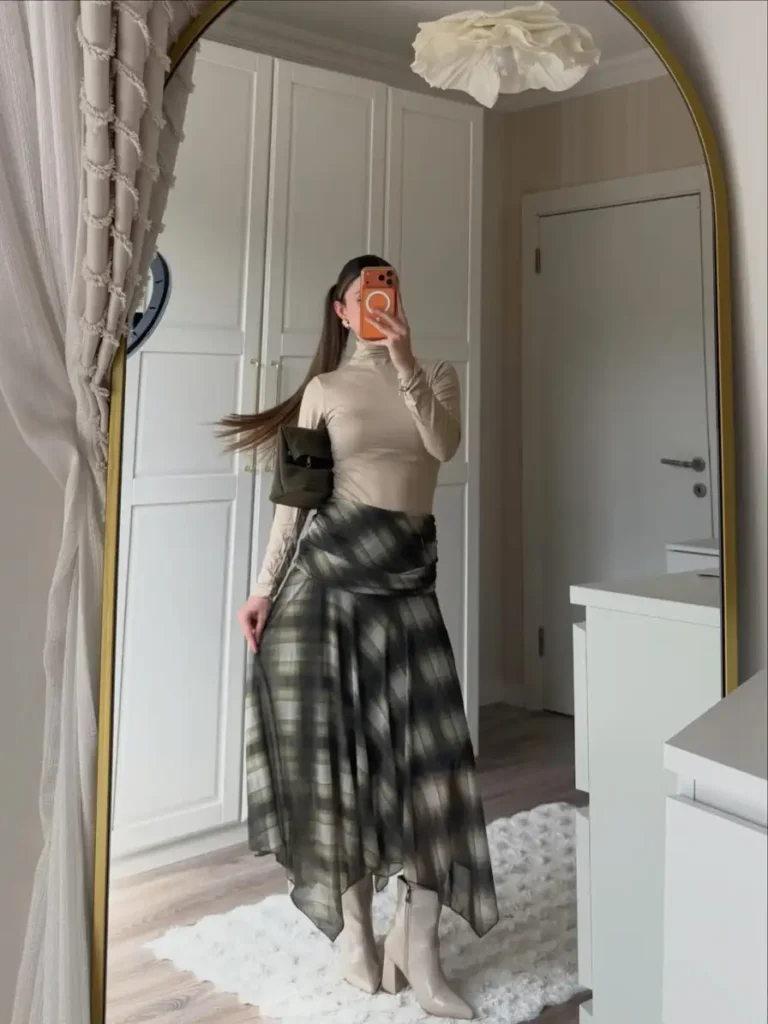 Woman wearing a fitted beige turtleneck with a green plaid draped midi skirt, cream ankle boots, and a dark handbag for a chic fall look.