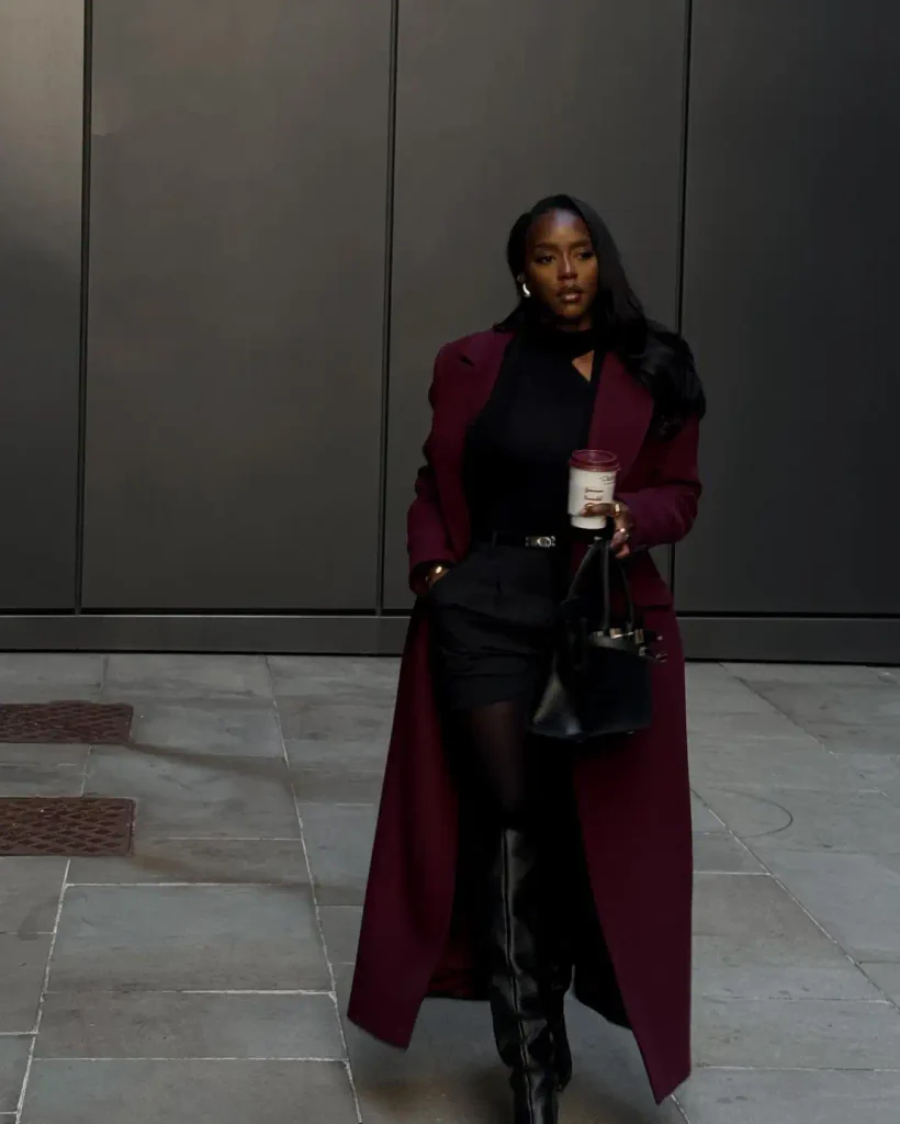  Black woman wearing a long burgundy coat over a black top and shorts with sheer black tights, tall leather boots, and a structured handbag.