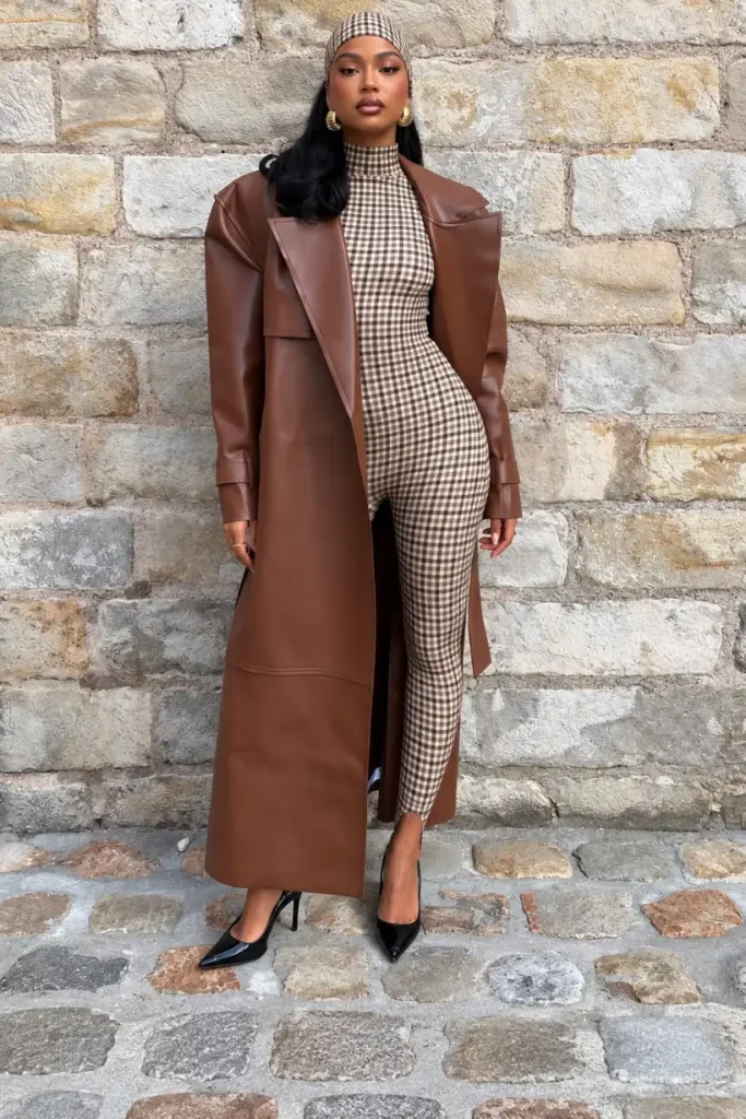 Fashionable Black woman wearing a chocolate brown leather trench coat over a fitted brown-and-cream gingham jumpsuit, with a matching checked headscarf and black pointed heels.