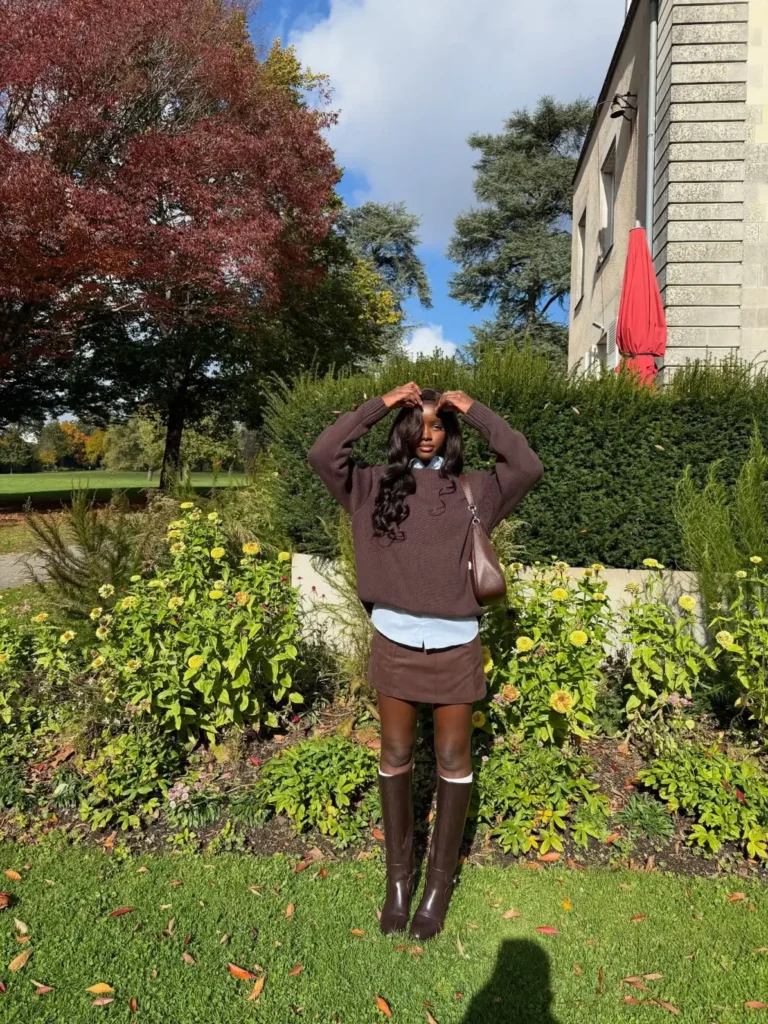 Black woman wearing an oversized chocolate brown sweater layered over a white shirt with a brown mini skirt, knee-high brown boots, and a shoulder bag for a preppy fall look.