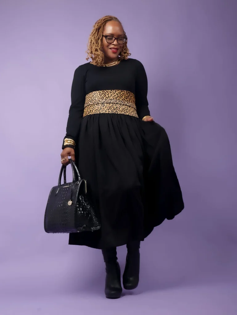 Black woman wearing a long black dress with a wide leopard-print waist detail, tall black boots, gold jewelry, and a structured black handbag for a classy fall look.