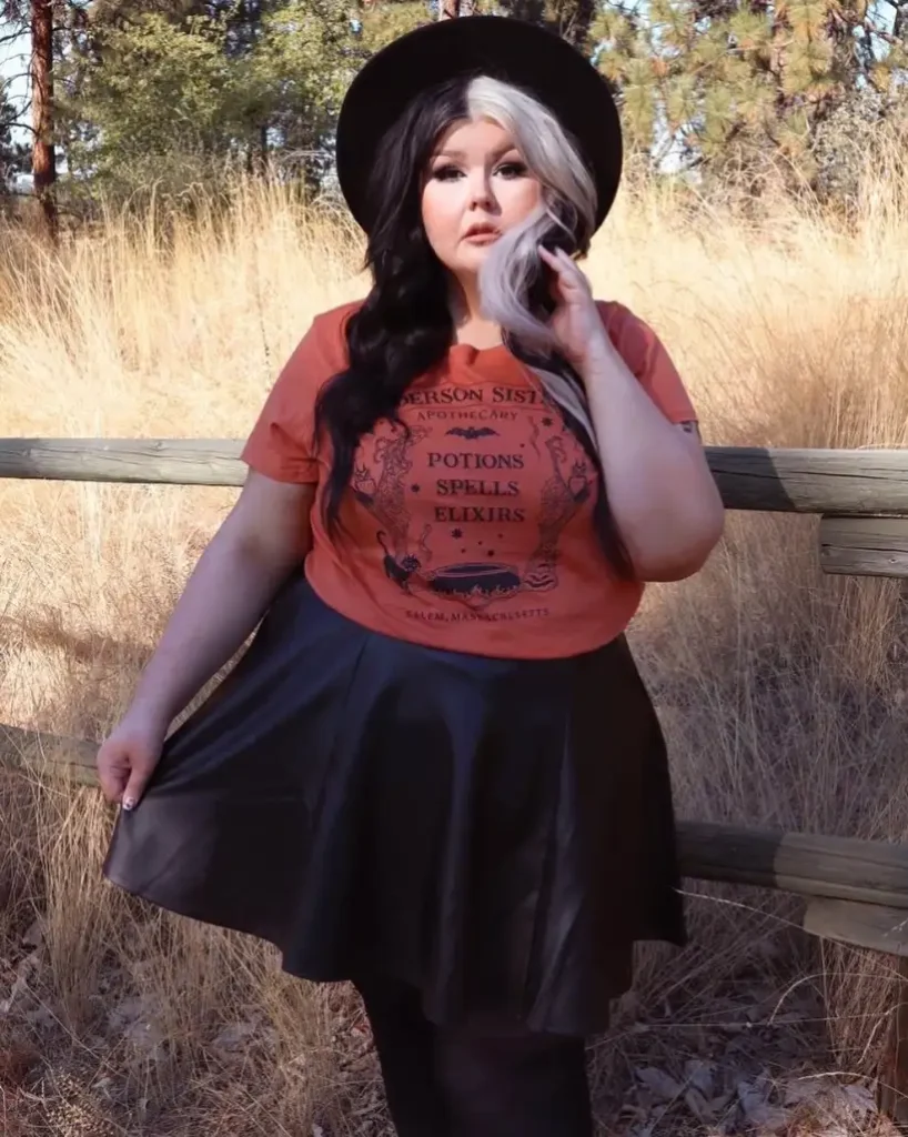 Plus size woman styling a casual witch Halloween outfit with an orange graphic tee, black skirt, and wide-brim hat.