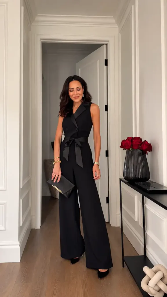 Woman wearing a classy sleeveless black wide-leg jumpsuit with double-breasted detailing, a tied waist, pointed black heels, and a matching clutch for a sophisticated New Year's Eve look.