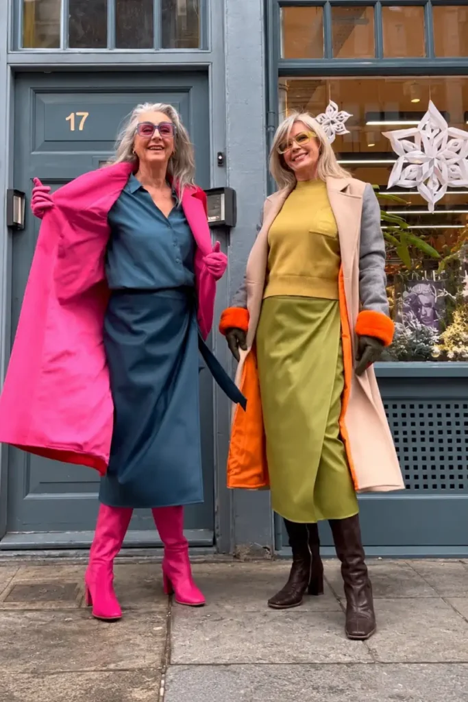 Two stylish women over 50 wearing colorful winter outfits featuring a hot-pink coat with teal dress and boots, and a cream coat with yellow knit and olive skirt.