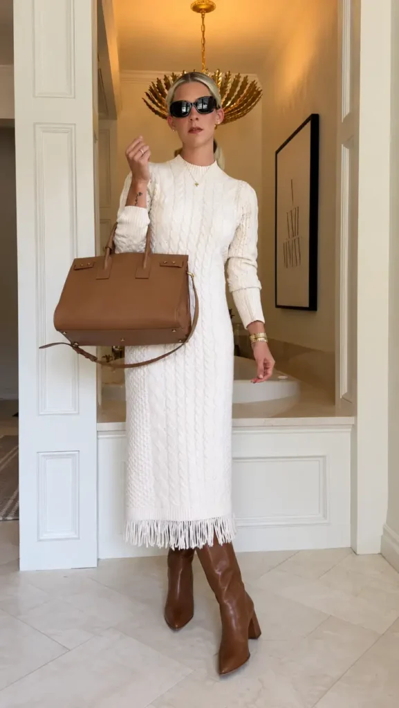 Woman wearing a cream cable-knit midi sweater dress with a fringed hem, cognac knee-high boots, and structured brown handbag for an elegant autumn outfit.