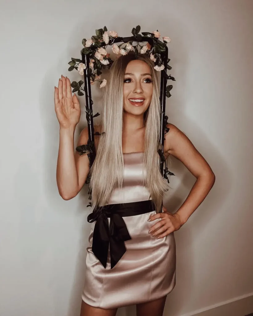 Woman wearing a pale satin mini dress with a black waist bow and a flower-covered frame around her head for a creative doll-inspired costume.