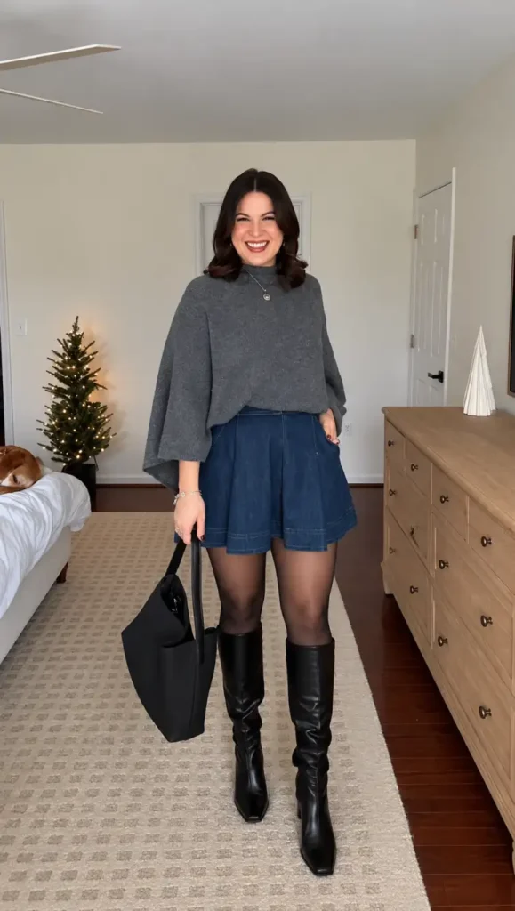 Woman wearing a loose gray cape-style sweater with a blue denim mini skirt, sheer black tights, knee-high black boots, and a black tote bag for a cozy fall look.