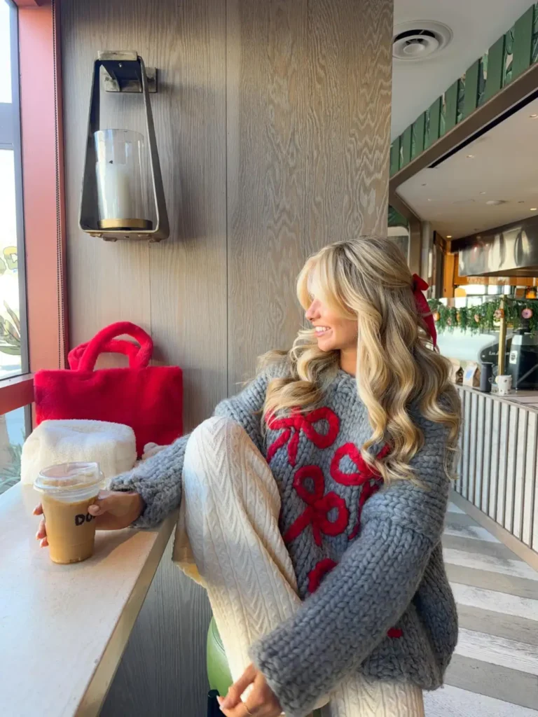 Woman wearing a chunky gray knit sweater with red bow details, cream cable knit trousers, and a fluffy red tote bag for a cozy casual Christmas outfit.