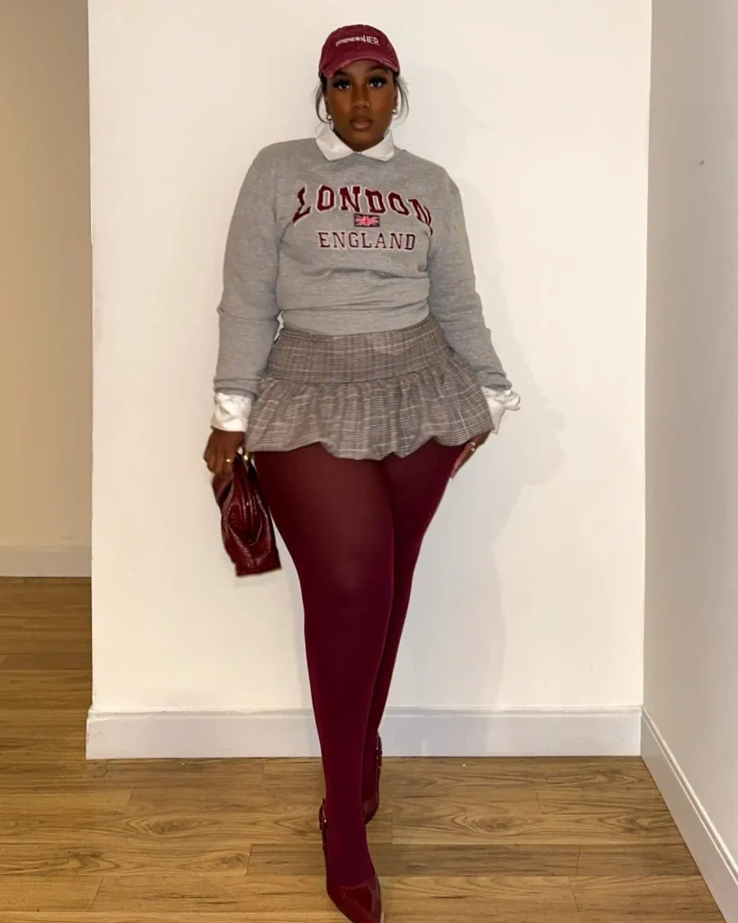 Curvy Black woman styling a gray London sweatshirt over a white collared shirt with a plaid mini skirt, burgundy tights, matching cap, and pointed shoes for a preppy fall look.