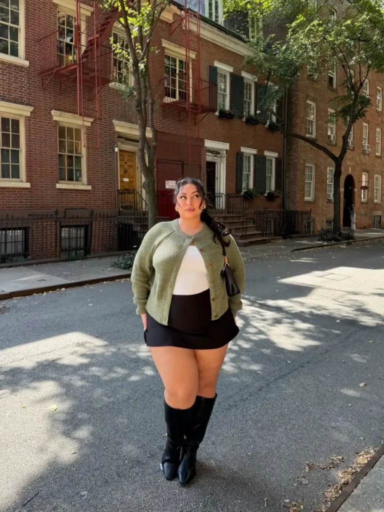 Plus size woman wearing a sage green cardigan over a white fitted top with a black mini skirt and tall black boots for fall street style.