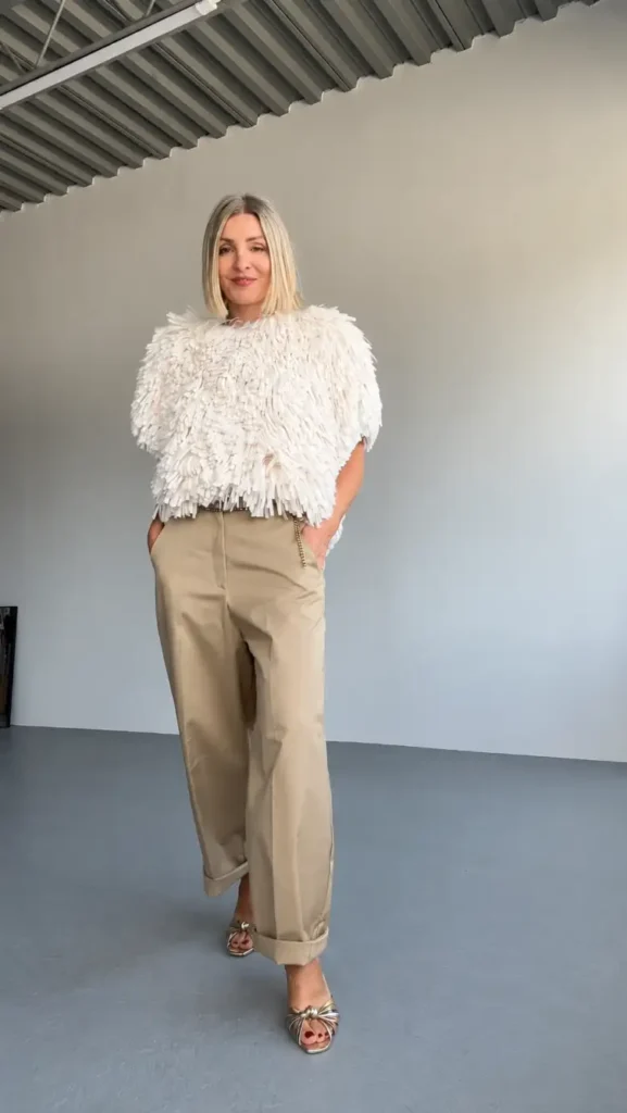 Stylish woman wearing a statement ivory textured top with beige cuffed trousers and metallic gold sandals for a sophisticated neutral Thanksgiving outfit.