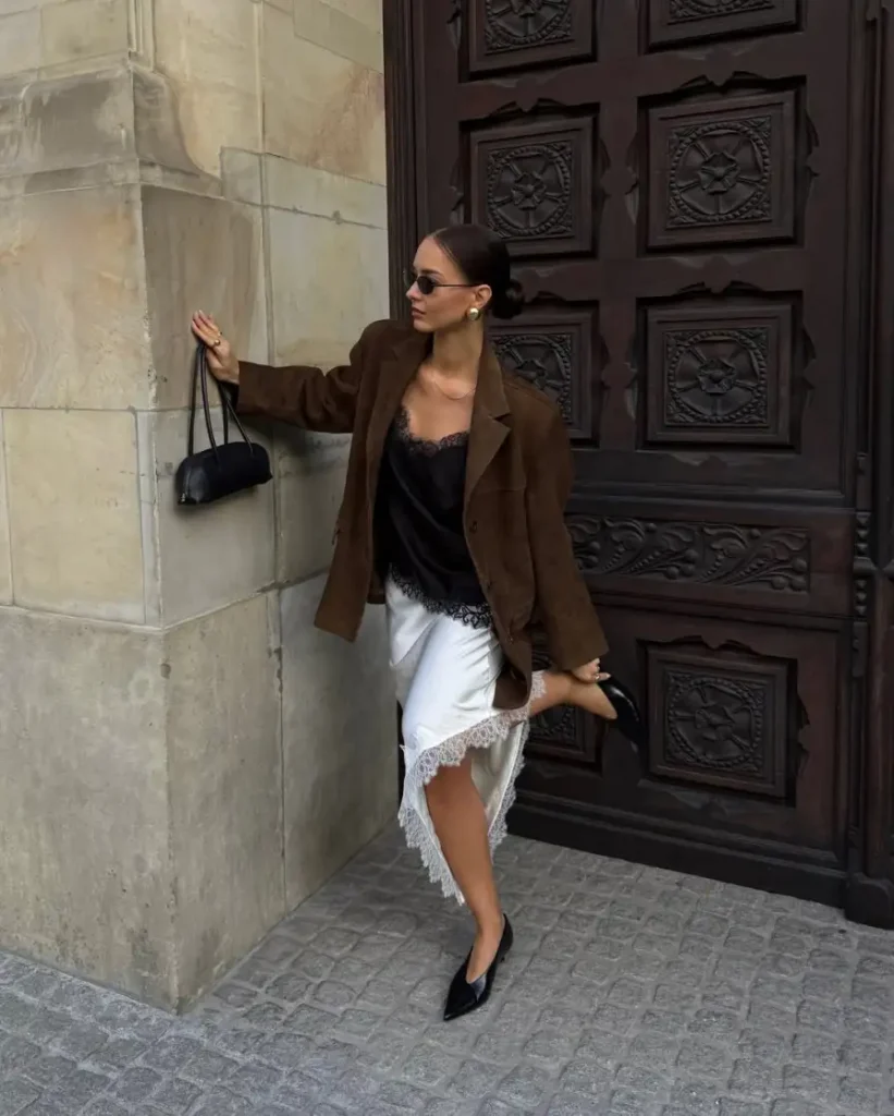 Woman wearing a brown blazer over a black lace camisole with a white satin lace-trim midi skirt and black pointed heels.