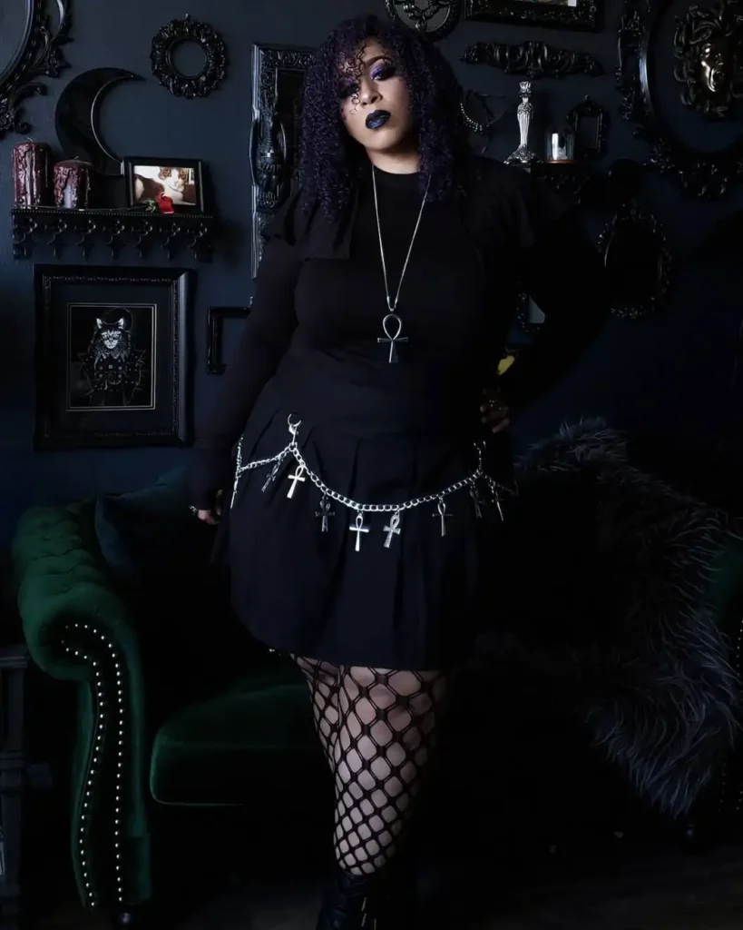 Plus size woman in an all-black goth Halloween outfit with a pleated skirt, fishnet tights, silver chains, and ankh jewelry.