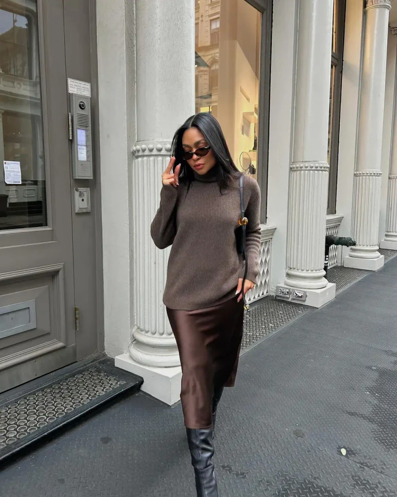 Black woman styling a taupe ribbed turtleneck sweater with a chocolate brown satin midi skirt, black knee-high boots, and narrow sunglasses.