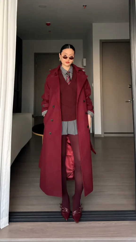 Woman wearing a long burgundy coat over a matching sweater vest, gray pinstripe shirt, striped tie, burgundy tights, and pointed shoes for a preppy winter fashion look.
