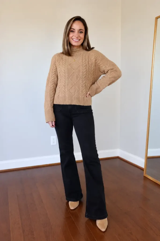 Woman wearing a camel cable-knit sweater with black flared jeans, tan pointed-toe ankle boots, and delicate gold jewelry for a cozy and classy Thanksgiving outfit.