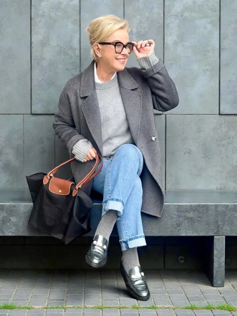 Chic woman over 50 wearing a charcoal-gray wool coat, gray sweater, light-wash cuffed jeans, gray socks, and black loafers for a casual winter street-style look.