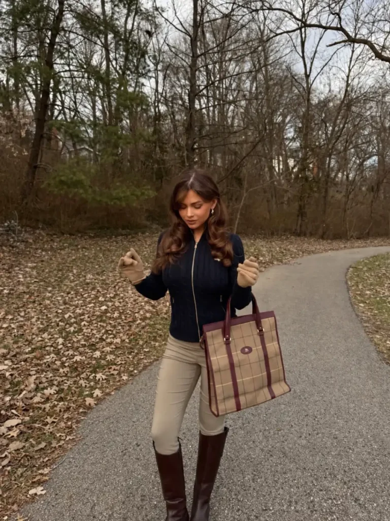 Woman wearing a navy zip-up knit with beige trousers, tall brown boots, gloves, and a plaid tote for an equestrian-inspired fall outfit.