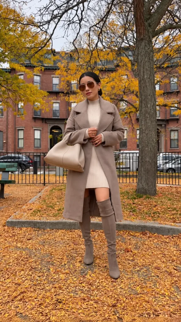 Petite woman wearing a taupe long coat over a cream ribbed mini sweater dress with taupe over-the-knee boots and a large neutral handbag.