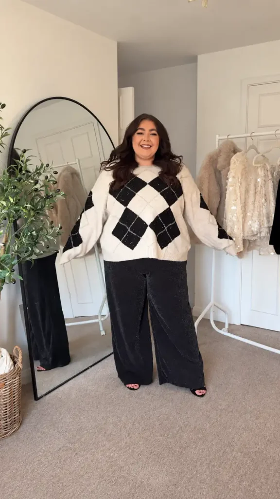 Plus-size woman pairing a cream and black argyle sweater with shimmering black wide-leg trousers and black open-toe shoes for a cozy Christmas party look.