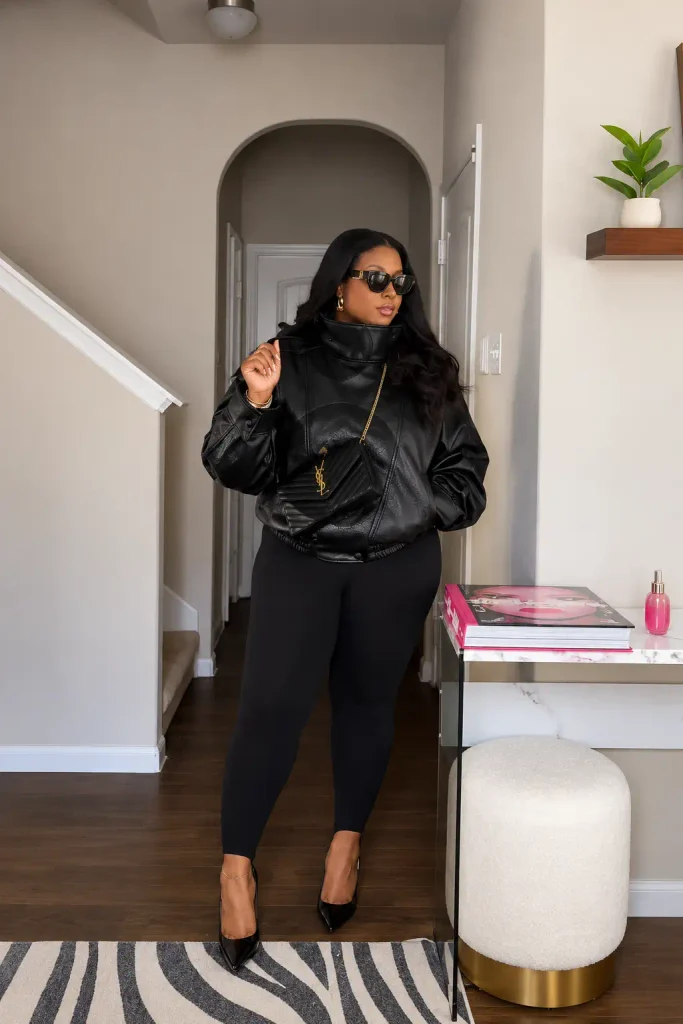 Plus size Black woman wearing an oversized black leather bomber jacket with fitted black leggings, pointed heels, sunglasses, and a quilted crossbody bag for an edgy fall outfit.