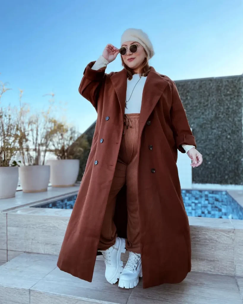 Plus size woman wearing a long rust-brown double-breasted coat with matching corduroy trousers, an ivory sweater, cream fluffy hat, and chunky white boots.