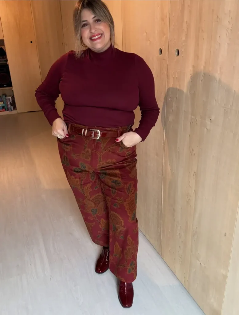 Plus size woman wearing a burgundy turtleneck with patterned wide-leg trousers, a brown belt, and glossy burgundy shoes for a retro fall look.