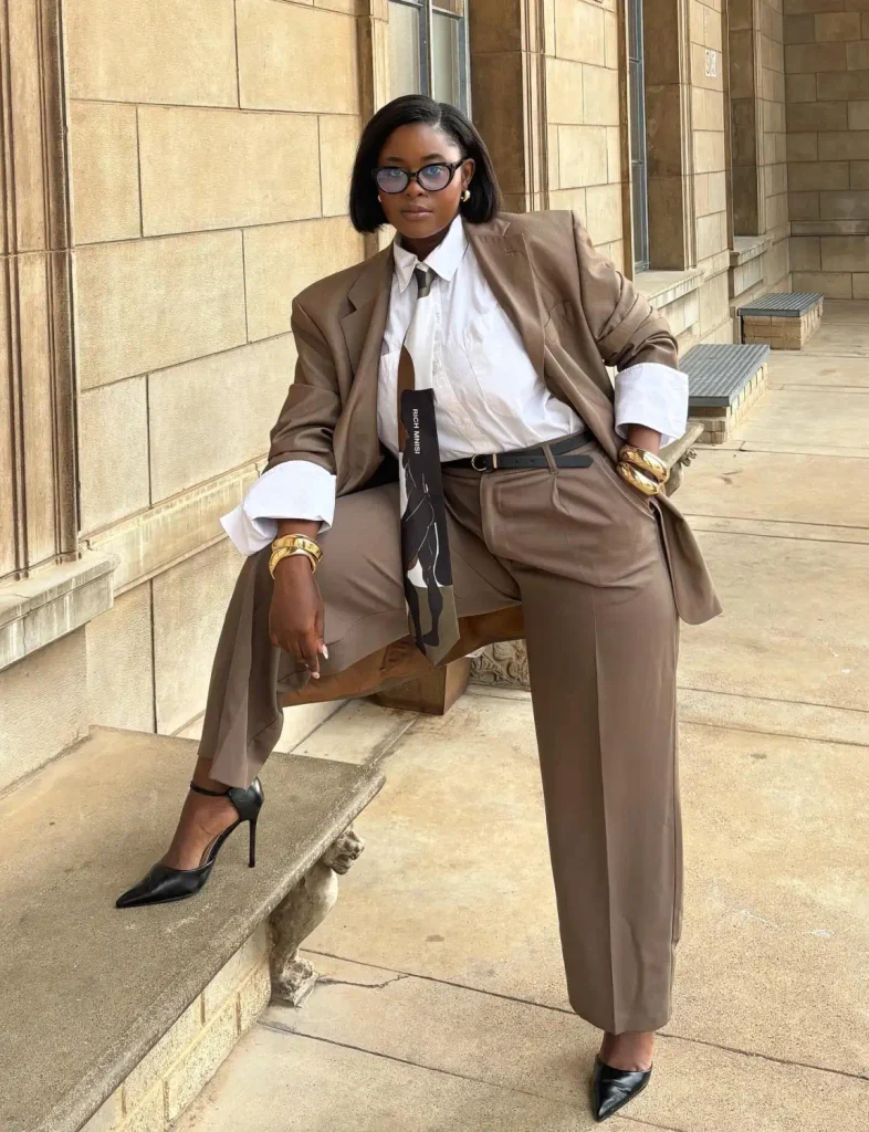 Black woman wearing an oversized taupe pantsuit with a white button-down shirt, patterned tie, black pointed heels, and gold bracelets for a powerful business outfit.