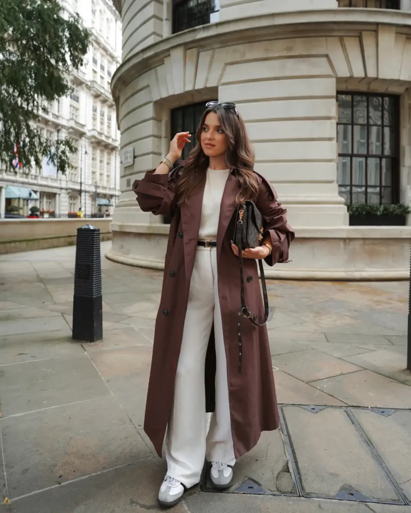 Long chocolate brown trench coat styled with a cream top, wide-leg white trousers, sneakers, and dark handbag for polished fall outfits.