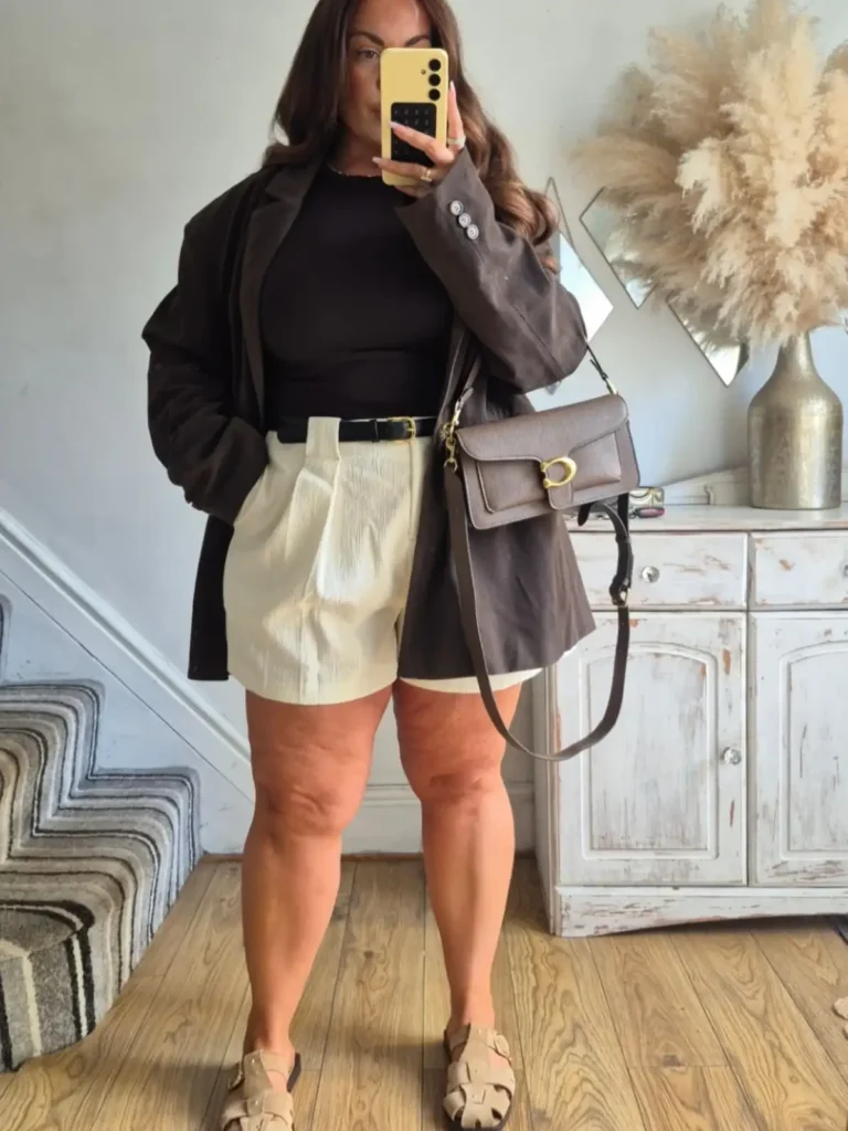 Plus size fall outfit with an oversized brown blazer, black fitted top, cream tailored shorts, black belt, neutral sandals, and brown shoulder bag.