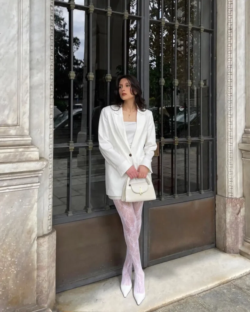 Woman wearing a white blazer with white floral lace tights, white pointed heels, and a structured white handbag.