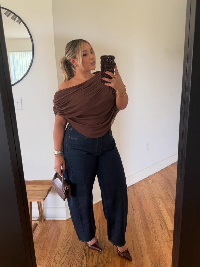 Woman wearing a chocolate brown draped one-shoulder top with dark-wash barrel-leg jeans, burgundy pointed-toe heels, and a structured brown handbag for an elegant fall dinner outfit.