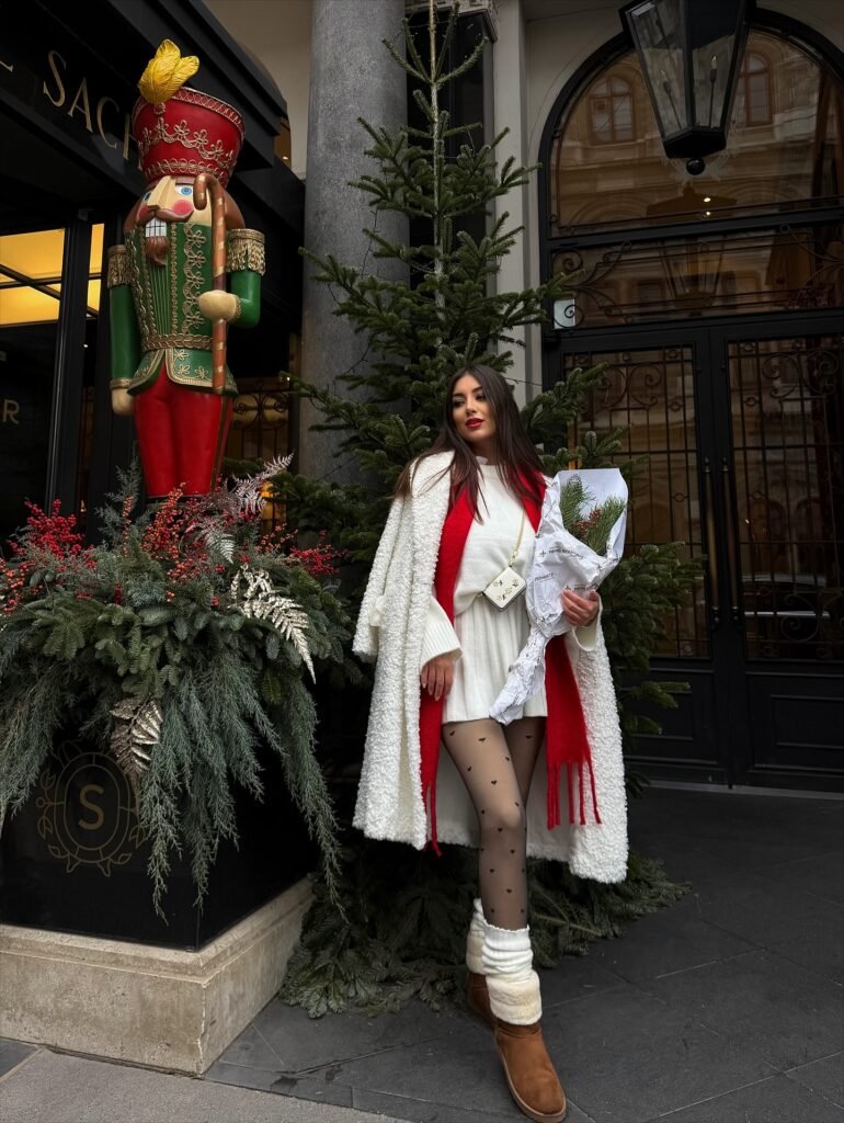 Woman wearing a white knit mini outfit with a fluffy white coat, bright red scarf, heart patterned tights, and brown winter boots for a cozy Christmas look.